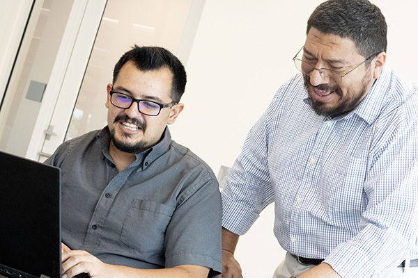 Male student in grey shirt examines research on his laptop with male mentor.