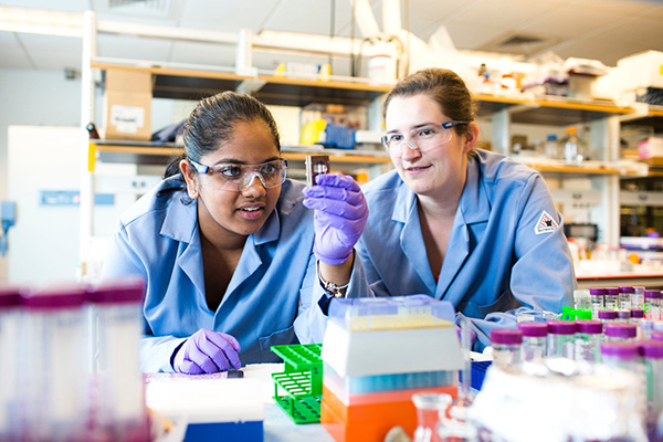 Two female students with blue lab coats and goggles work diligently in the lab.