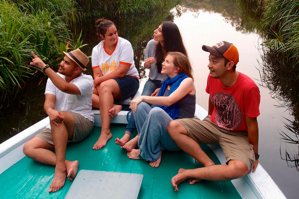 Five people sitting barefoot on a small boat in a narrow waterway, smiling and talking together while one person points toward the distance.